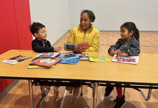 An adult and two children sit at a table reading books in a gymnasium.