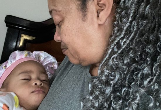 Woman with gray braids holding a sleeping baby in animal pajamas and a bonnet, sharing a tender moment.
