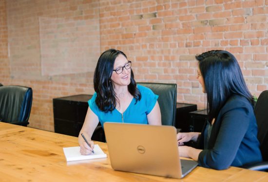 Two women sit at a conference table with a laptop, smiling and having a conversation.