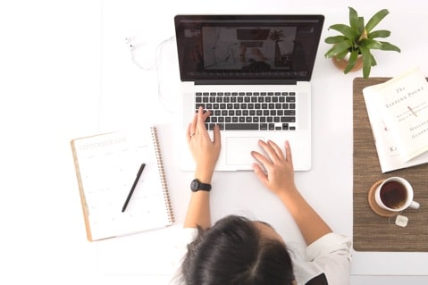 Person working on a laptop at a white desk with a planner, plant, book, and cup of coffee.