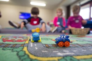 Two toy cars on a play mat with children sitting and playing in the blurred background.