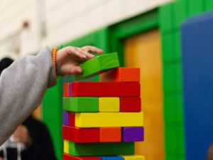 A hand placing a green block on top of a colorful wooden block tower indoors.