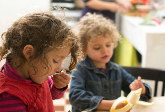 Two young children eat fruit at a table while an adult prepares food in the background kitchen.