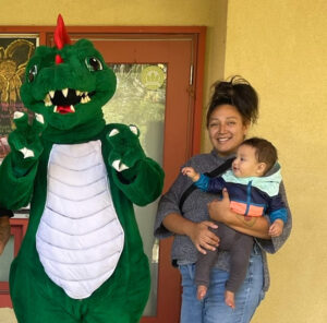 A woman holding a baby stands next to a person in a green dragon costume, all smiling outdoors.