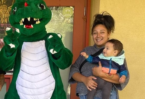 A woman holding a baby stands next to a person in a green dragon costume, all smiling outdoors.