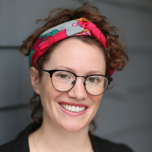 Smiling person with curly hair, glasses, and a colorful headband, standing in front of a gray wall.