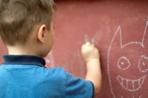 A young boy draws with white chalk on a red wall, creating a smiling face with pointy ears.
