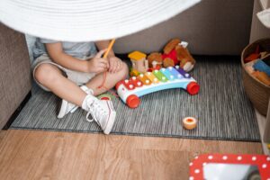 Child sitting on rug playing with xylophone, surrounded by toys and wearing white sneakers under a white canopy.