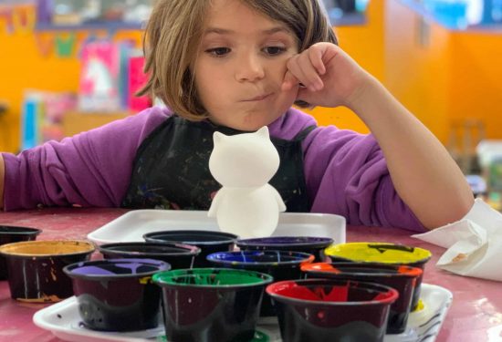 A child sits at a table, thoughtfully looking at a white ceramic cat and several paint cups.