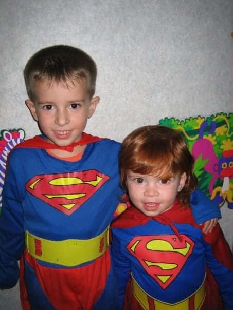 Two young children in Superman costumes stand together, smiling in front of a colorful wall with animal art.