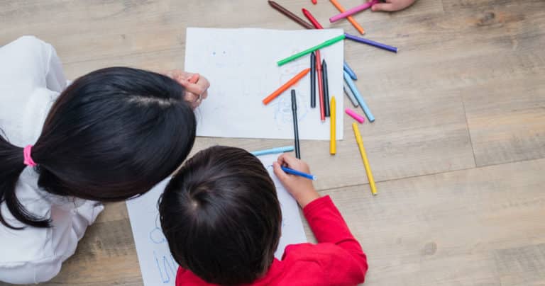 Two children drawing with colored markers on white paper, seen from above on a wooden floor.