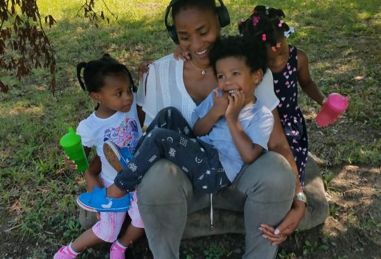 A woman with headphones sits on a rock outdoors, smiling with three young children holding colorful cups.