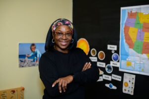 Smiling woman with glasses and headband stands with folded arms in a classroom with maps and space posters.