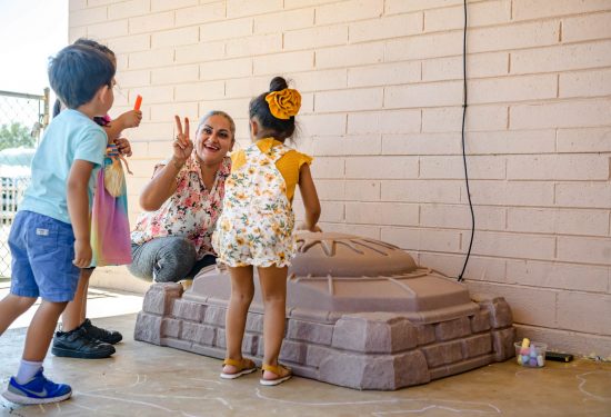 Woman smiling and flashing a peace sign as two children play with chalk near a sandbox outside.