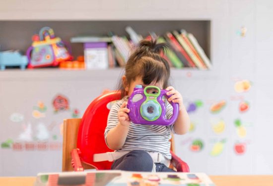 A young child in a red chair holds a toy camera up to their face, with books and toys in the background.