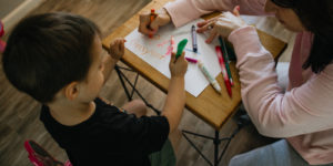Child and adult drawing with markers at a small wooden table, creating colorful artwork together.