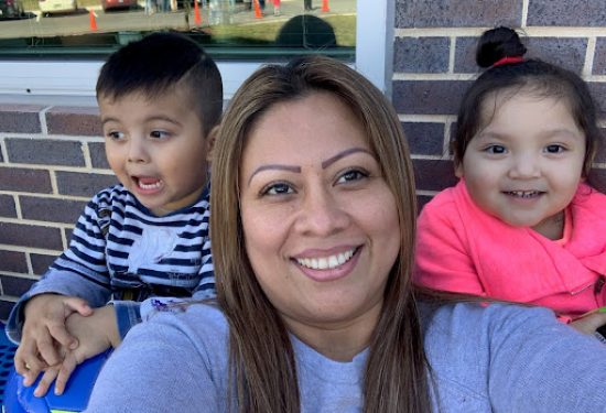 A woman smiles for a selfie with two young children in front of a brick wall.