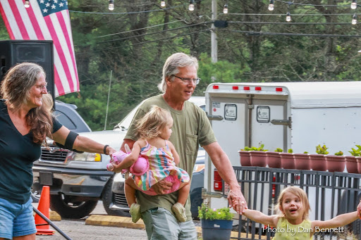 A man and woman walk hand-in-hand with two young girls at an outdoor event, American flag in background.