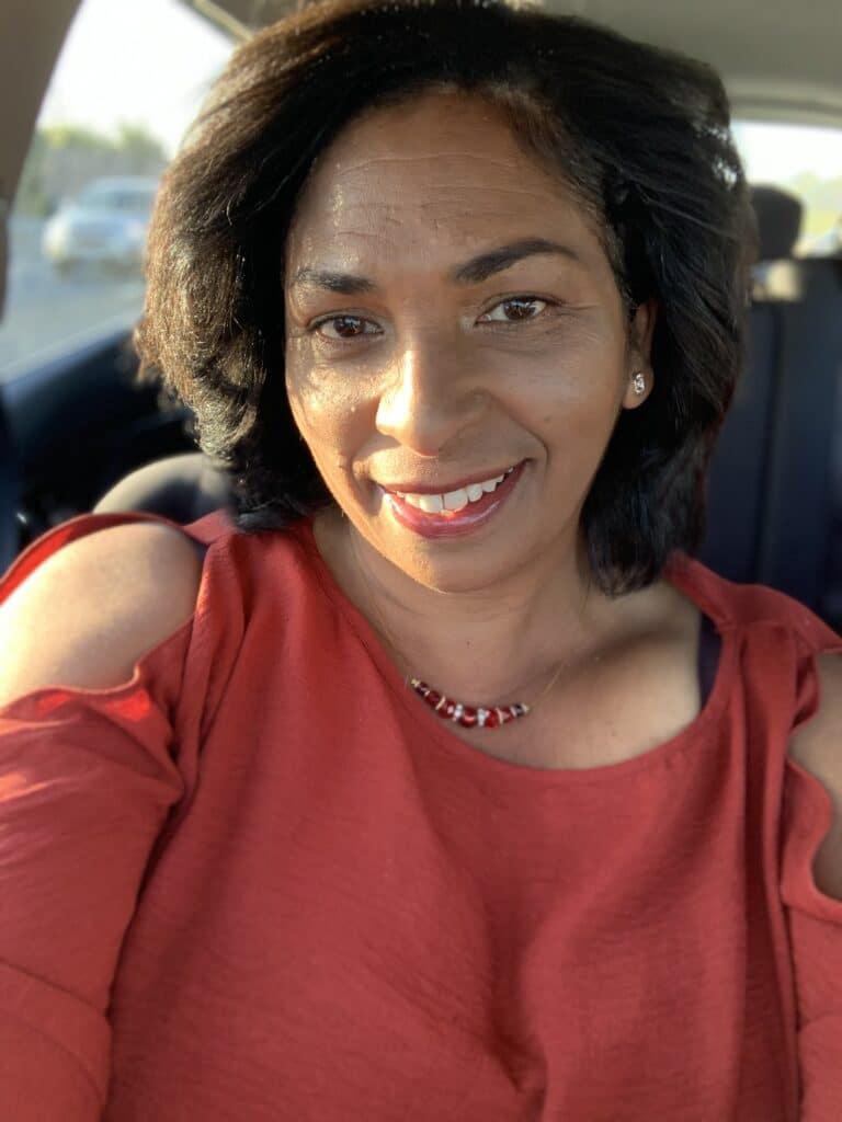 Woman with dark hair wearing a red top smiles while sitting in a car, sunlight on her face.