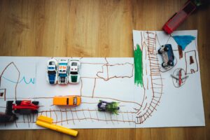 Toy cars on a hand-drawn road map made of paper, placed on a wooden floor with two yellow markers nearby.