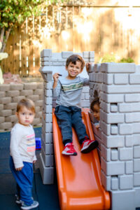 Three young children play on a plastic playset with a slide in a sunlit backyard.