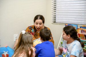 Woman reading a book to three children, all engaged and smiling, in a cozy room with bookshelves.