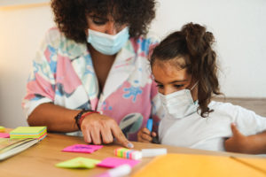 Adult and child wearing masks, sitting at a table with colorful sticky notes and markers, working together.