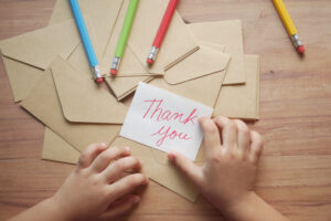 A child’s hands hold a “Thank you” card above brown envelopes and colored pencils on a wooden table.