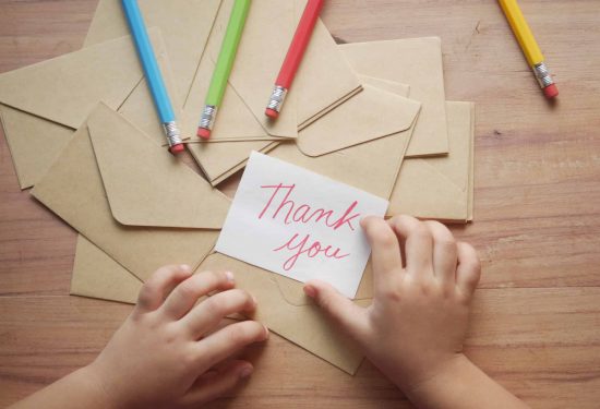 Child's hands holding a "Thank you" card above brown envelopes and colored pencils on a wooden table.