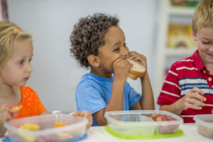 Three young children sit at a table eating lunch from plastic containers, smiling and enjoying their food.