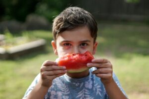 Young boy holding and eating a slice of watermelon outdoors, looking directly at the camera.