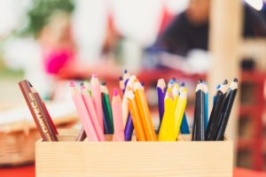 A wooden box holds colorful pencils, including pink, yellow, blue, and black, on a blurred background.