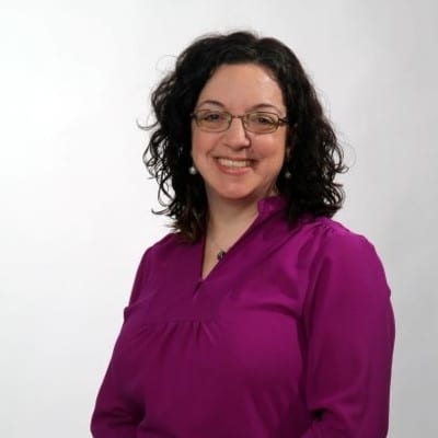 Woman with curly dark hair and glasses, wearing a purple blouse, smiles in front of a plain light background.