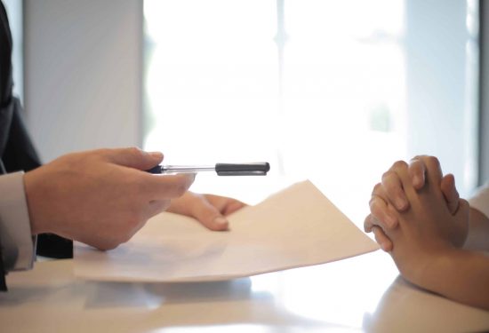 Person handing a pen and document to another person whose hands are clasped on a table.