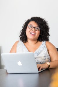 Smiling woman with curly hair and glasses working on a laptop at a desk.