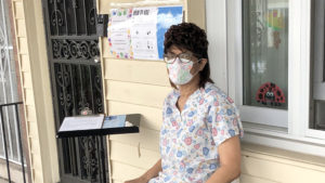 Woman in scrubs and floral mask sits outside a house near a window with notices and a ladybug decoration.