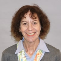 Woman with curly brown hair, wearing a gray blazer, light blue shirt, and floral scarf, smiling at camera.