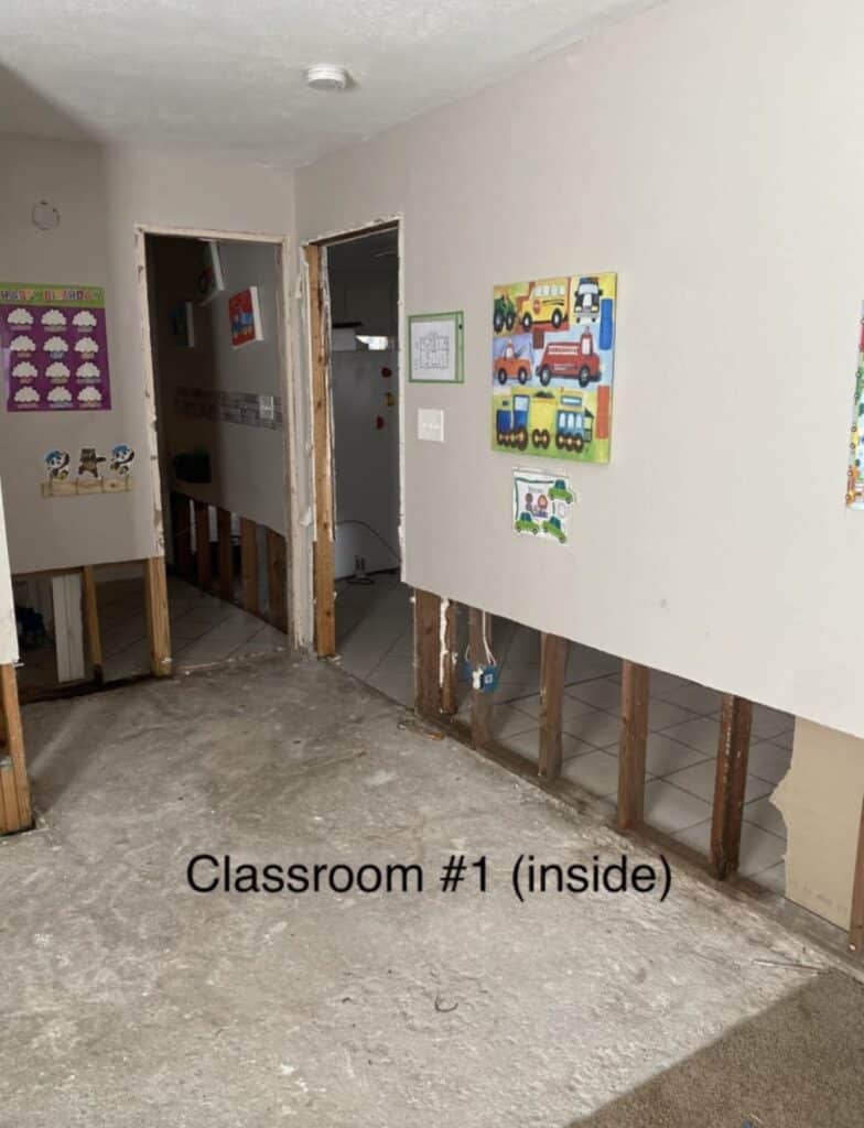 A classroom with damaged lower walls, exposed studs, bare floor, and children's artwork on the walls.