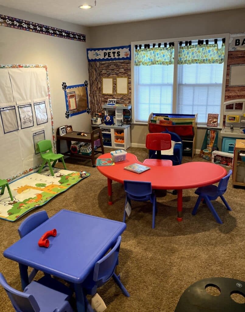 Colorful preschool classroom with small tables, chairs, toys, play mat, and learning centers by the windows.