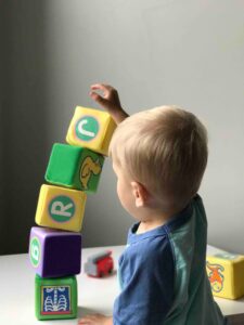 Toddler stacking colorful toy blocks on a white table, with a small toy train in the background.