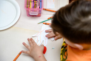 A child colors a paper hand cutout with crayons at a table.