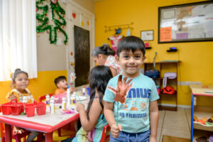 Smiling boy with painted hand stands in classroom; other children and teacher work at tables in the background.