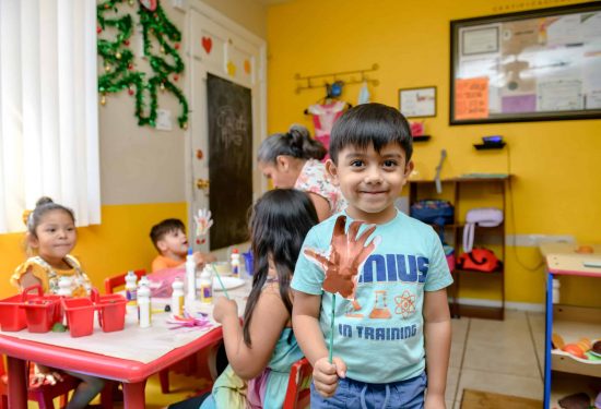Smiling boy with painted hand stands in classroom; other children and teacher work at tables in the background.