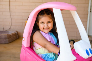 Smiling young girl in a rainbow dress sitting in a pink toy car outdoors.