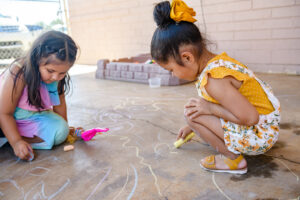 Two young girls drawing colorful chalk art on a concrete sidewalk outside on a sunny day.