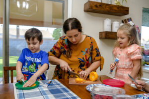 A woman and two children cutting fruit together at a kitchen table, smiling and engaged.
