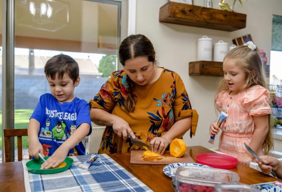 A woman and two children cut fruit together at a kitchen table with plates and snacks.
