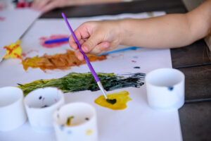 A child’s hand uses a paintbrush to mix colors on paper, with paint cups nearby on a table.