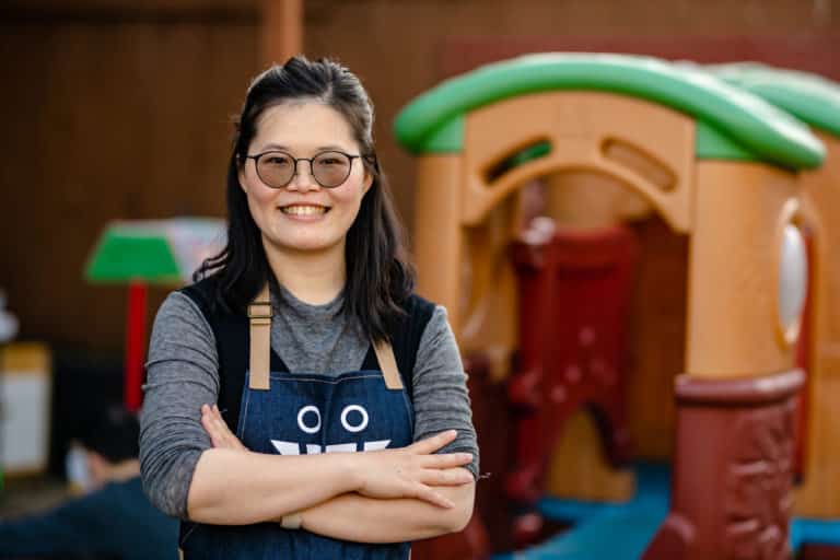Smiling woman with glasses and apron stands with arms crossed in front of colorful playground equipment.