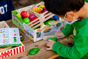 Child playing with toy fruits and a wooden knife at a table, with boxes of play food nearby.